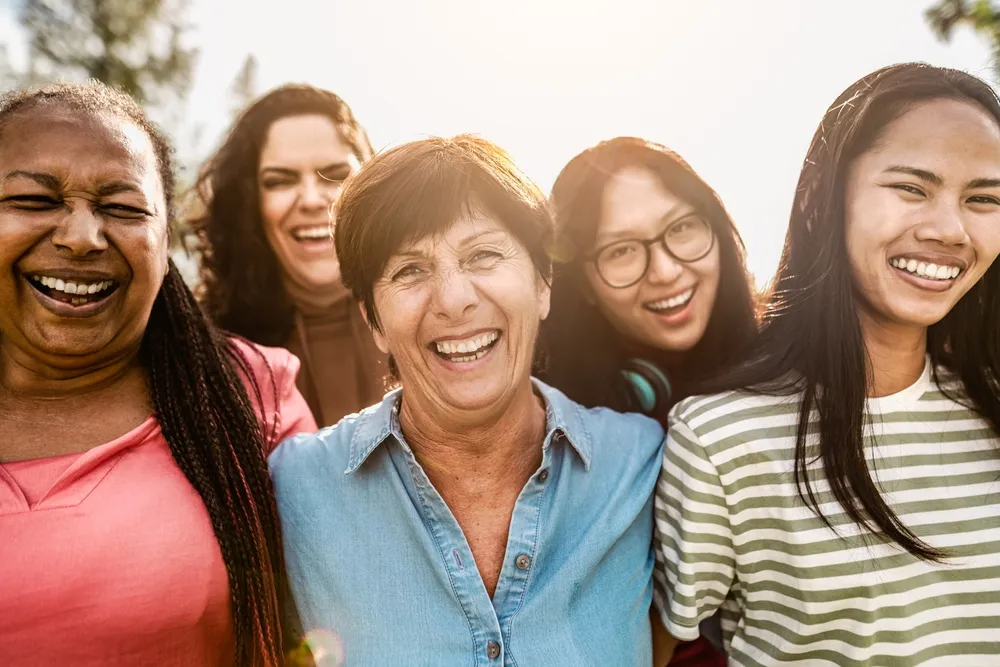 Five diverse women smiling and laughing together outdoors in sunlight.