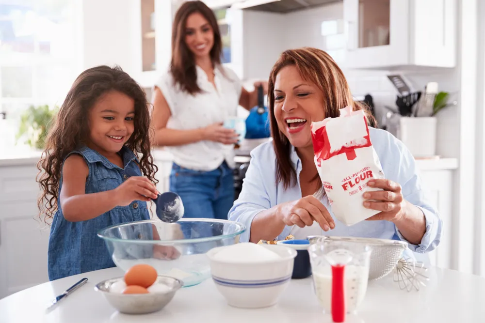 Smiling girl and woman baking with flour and ingredients in a bright kitchen, another woman watching.