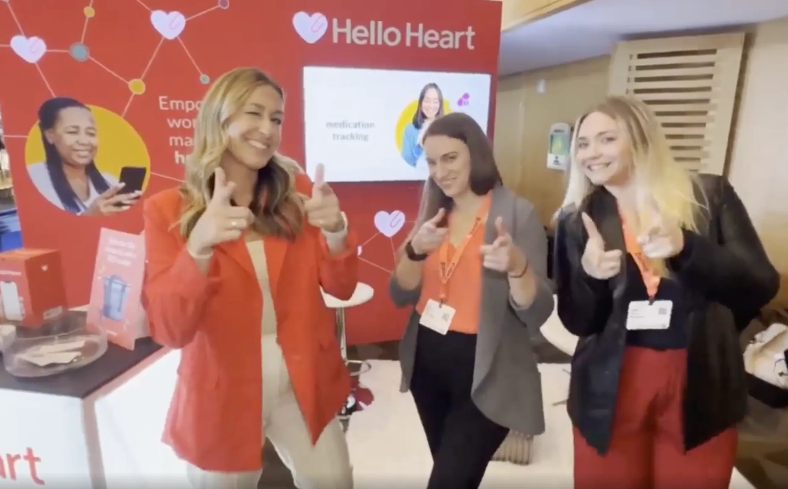 Three smiling women posing with finger guns in front of a Hello Heart event backdrop.