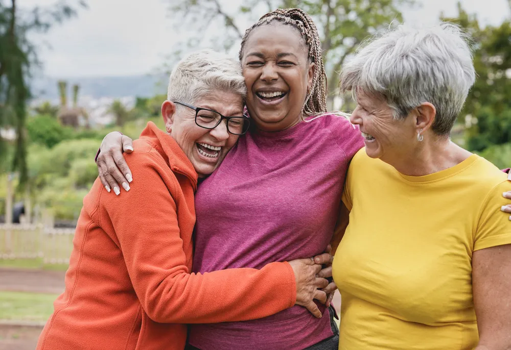 Three joyful women hugging and laughing outdoors in colorful casual clothes.