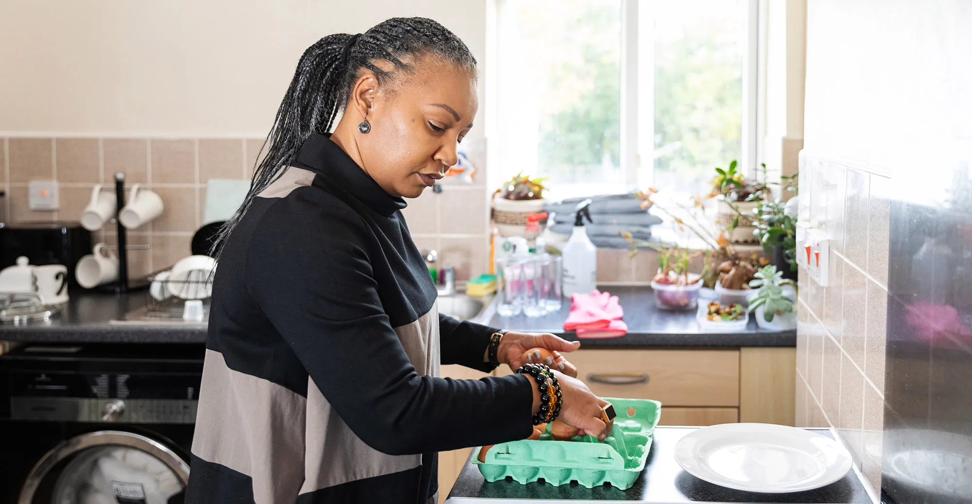 Woman cracking an egg into a green carton in a bright kitchen with plants and dishes on the counter.