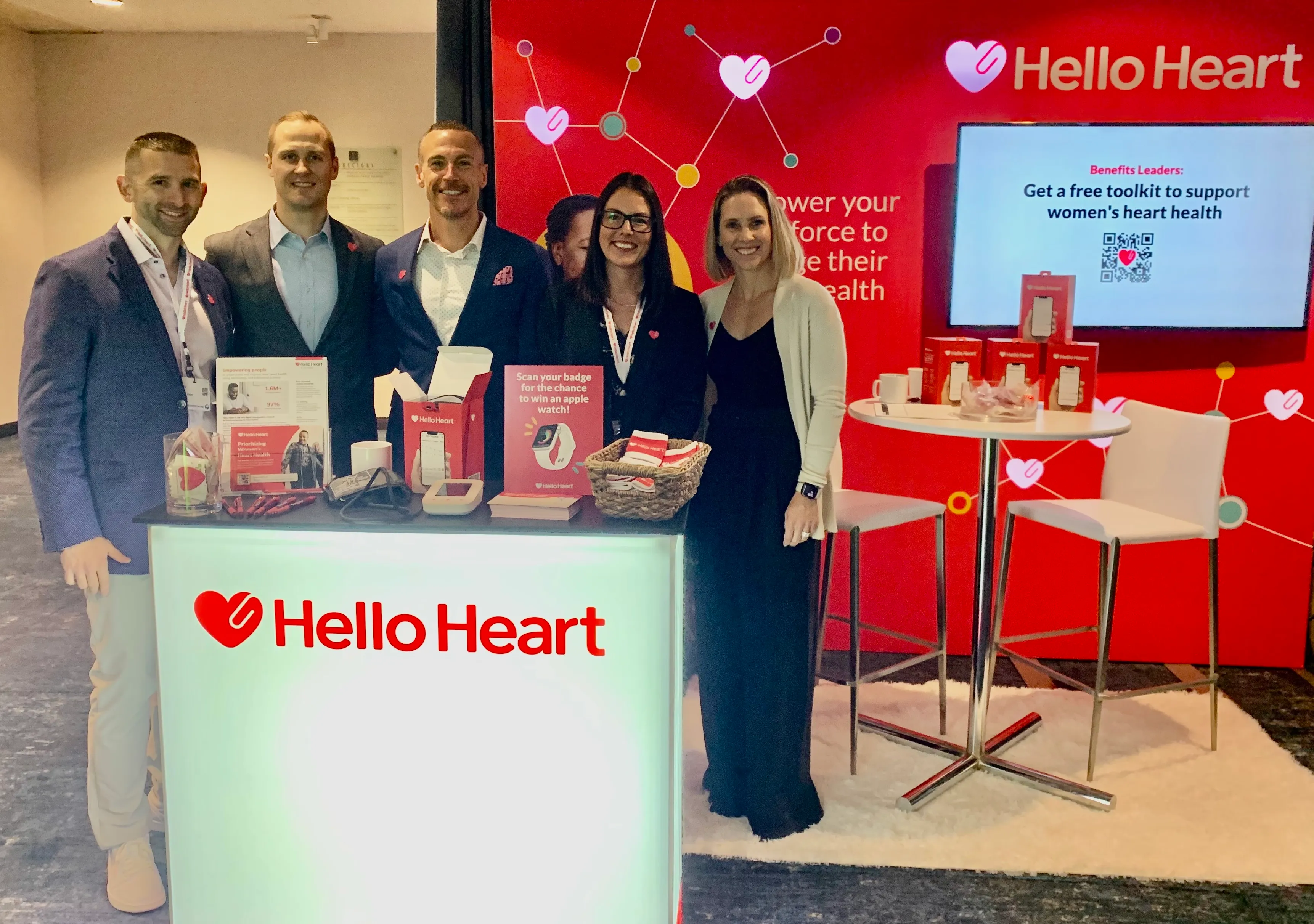 Five people smiling at a Hello Heart exhibit booth with heart health info and promo materials.