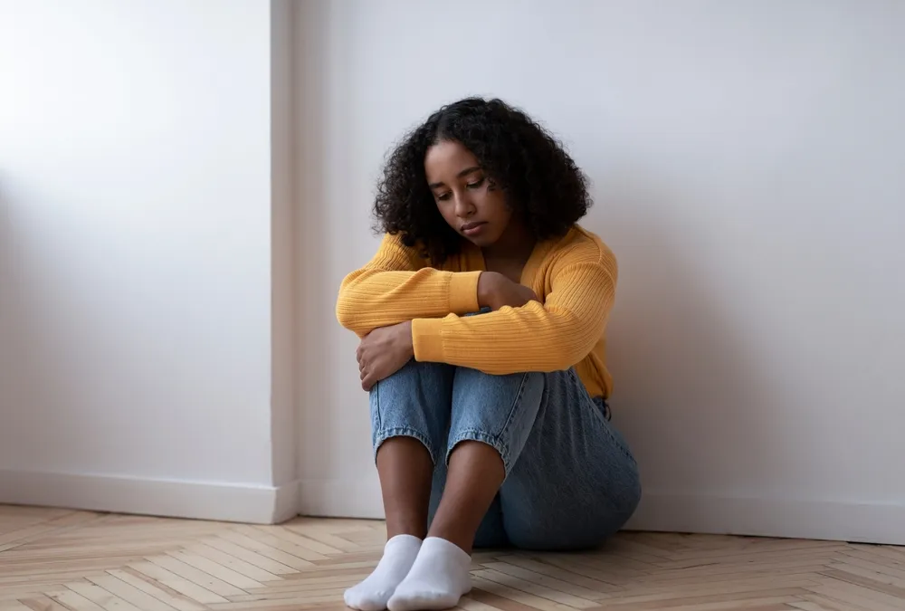 Woman in yellow sweater sitting on wooden floor against white wall, hugging knees and looking down.