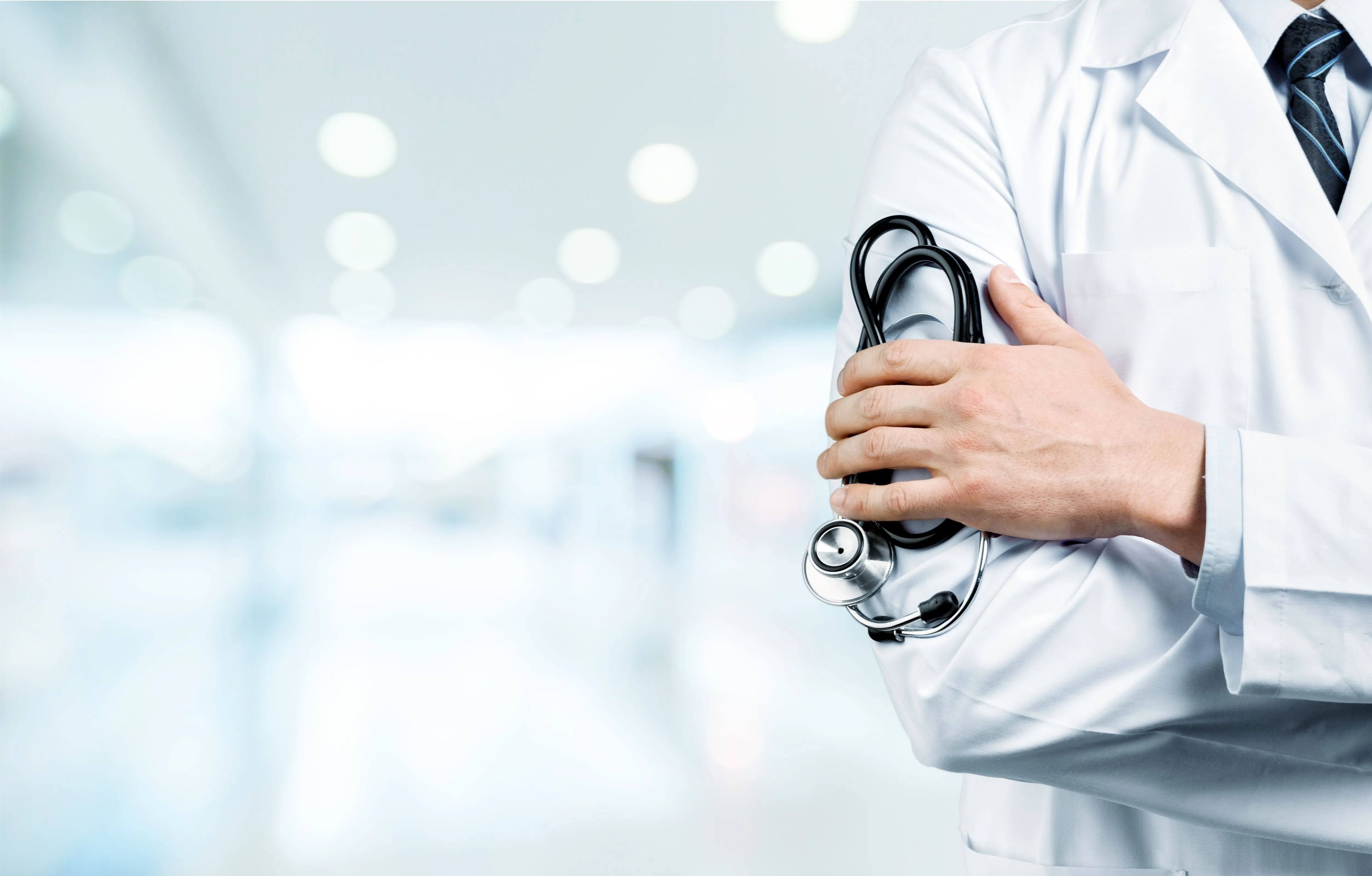 Doctor in white coat with striped tie holding a stethoscope, blurred hospital background.