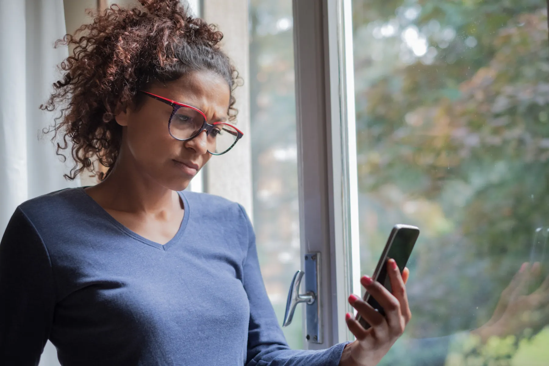 Woman with curly hair and glasses looking at her smartphone with a concerned expression by a window.