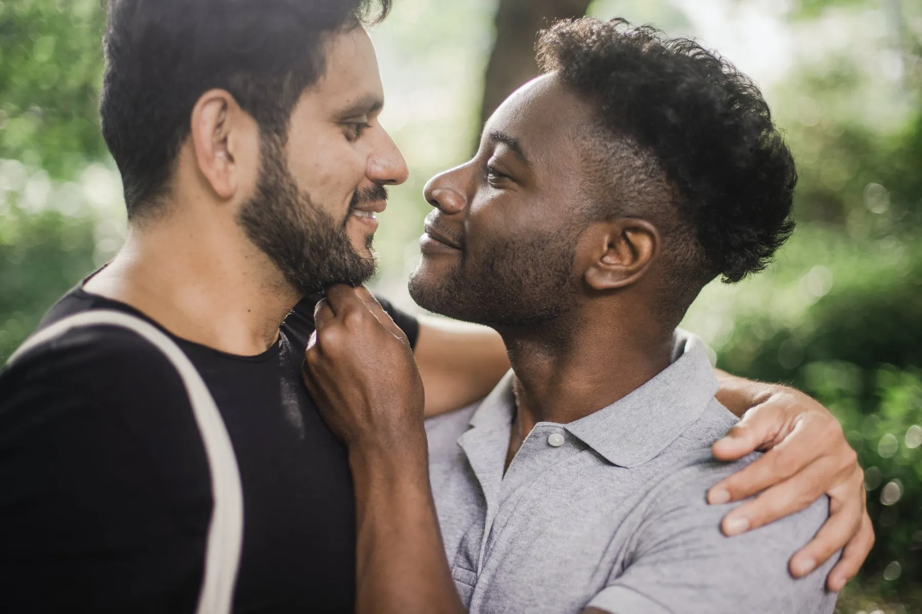 Two men smiling and embracing closely in an outdoor setting with greenery behind them.