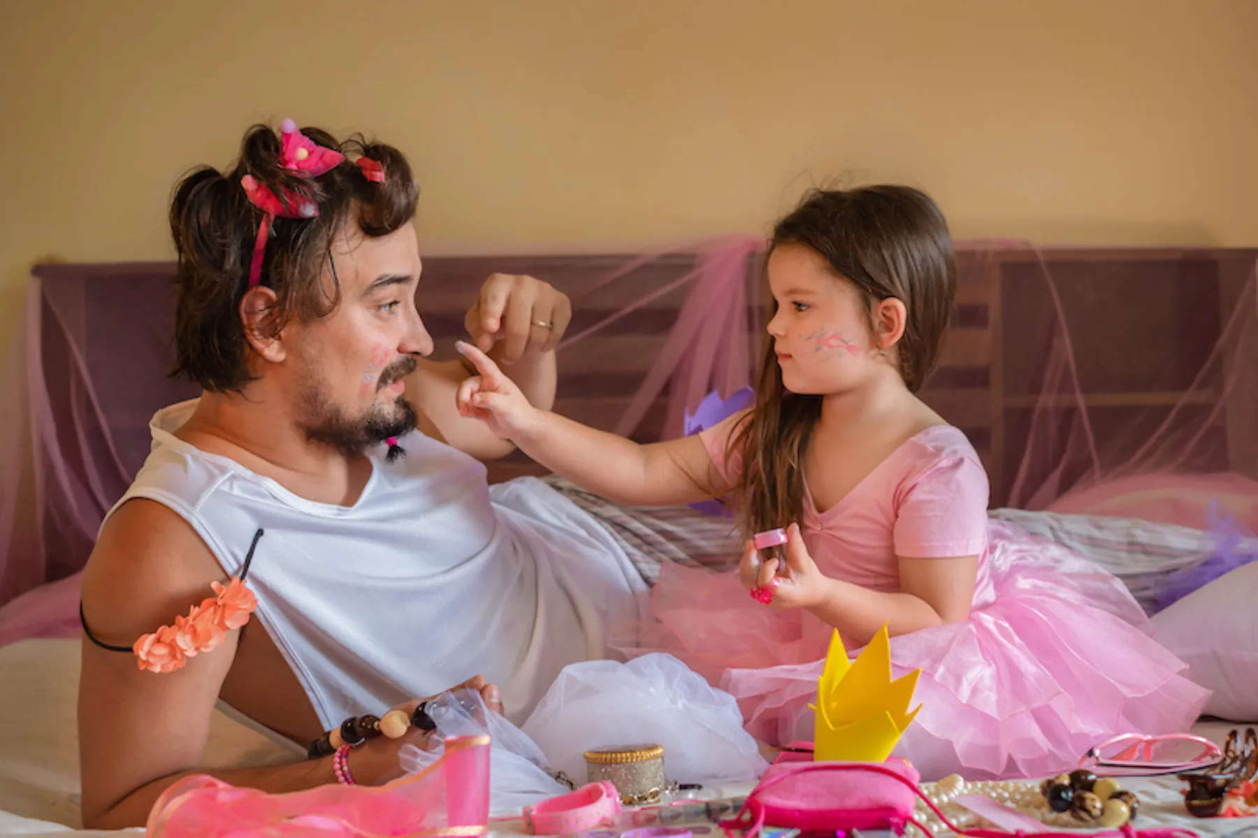 Father and daughter in dress-up clothes play face painting together on a bed with accessories.