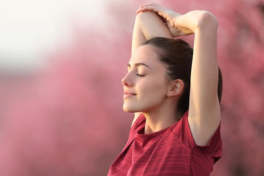 Woman in red shirt stretching arms behind head with closed eyes against soft pink background.