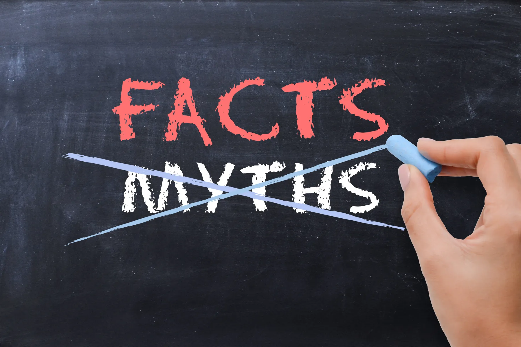 Hand crossing out the word MYTHS beneath the word FACTS on a black chalkboard with blue chalk.