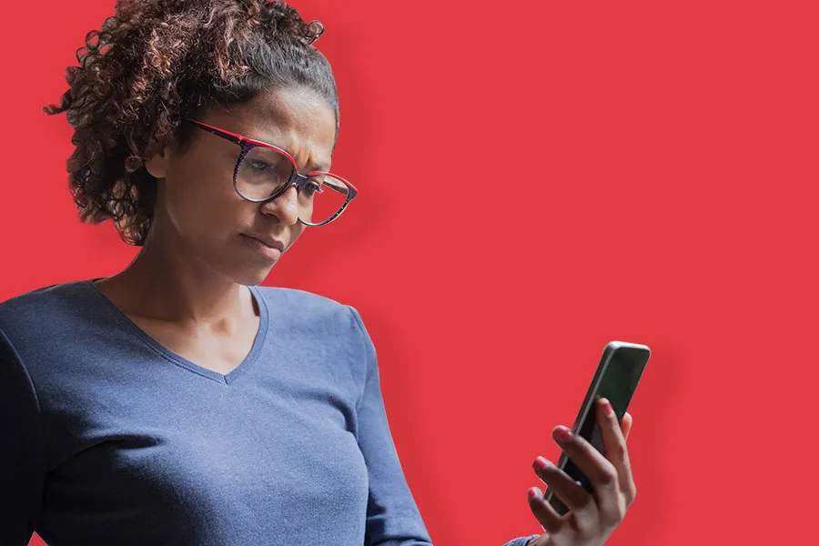 Woman with curly hair and glasses looking at phone with concerned expression on red background.