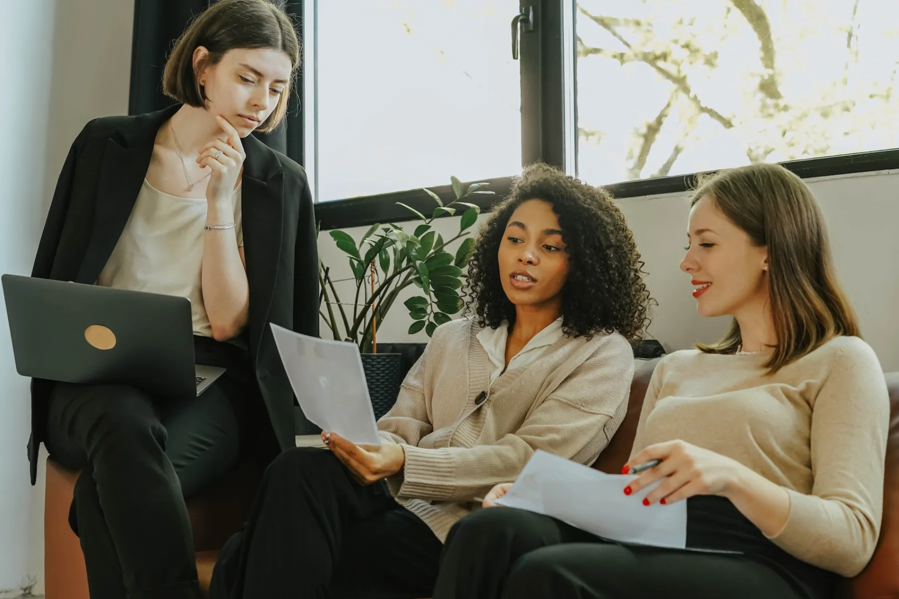 Three women discussing documents in a modern office with a laptop and plants by the window.