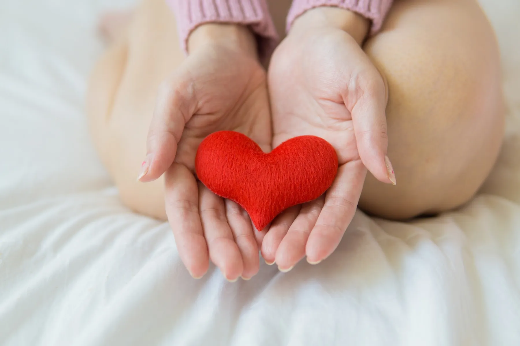 Hands gently holding a soft red heart-shaped object on a white bed with knees visible in background.