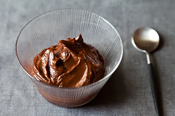 Glass bowl with thick chocolate mousse on a gray surface next to a spoon.