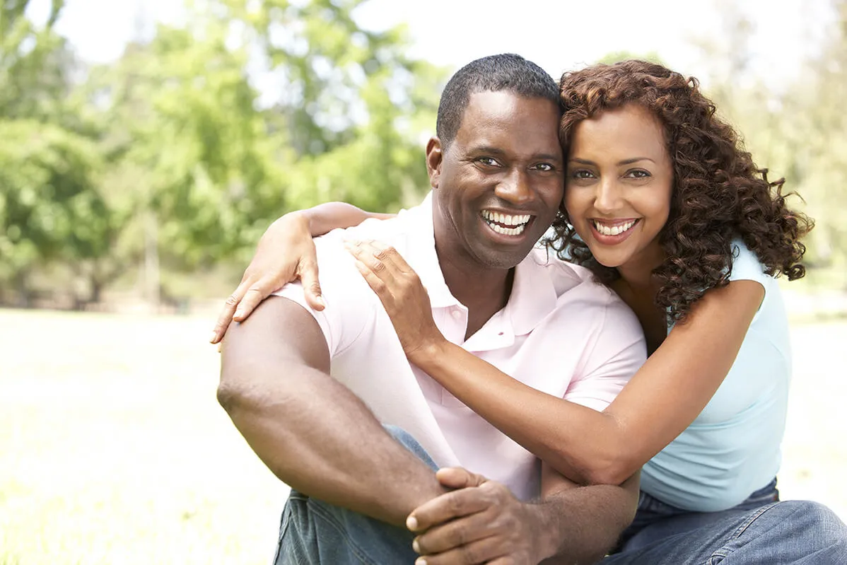 Smiling couple sitting on grass outdoors, the woman hugging the man from the side.