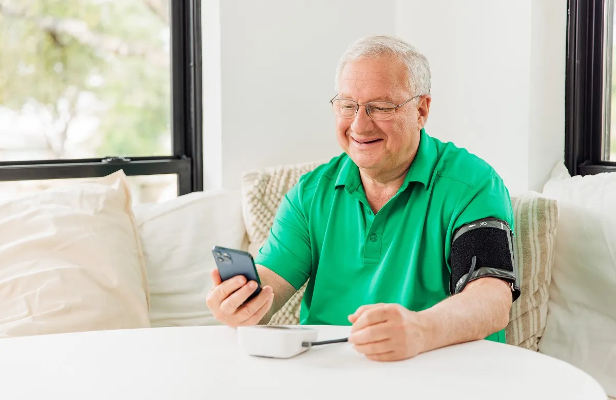 Older man in green shirt using a digital blood pressure monitor while looking at his phone.
