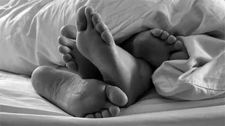 Close-up of four bare feet intertwined under white bed sheets in black and white photo.