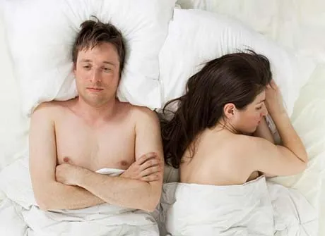 Man lying on bed with arms crossed looking away while woman sleeps on her side facing away.