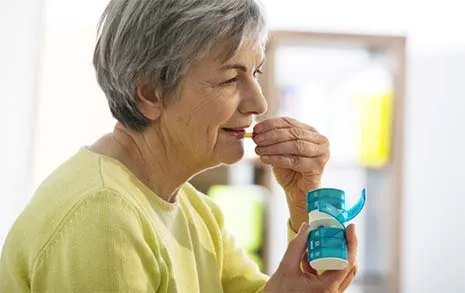 Older woman taking medication from a blue pill organizer in a bright room.