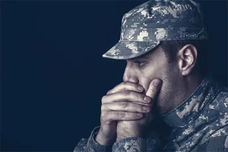 Pensive soldier in camouflage uniform resting his face on clasped hands against dark background.
