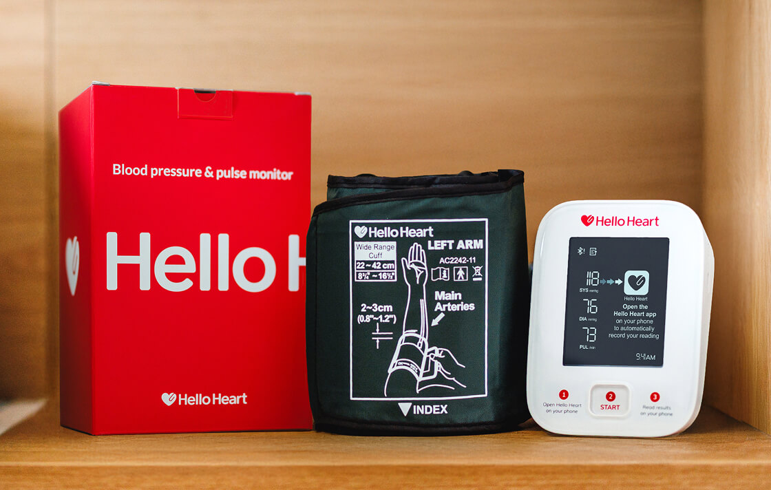 Hello Heart blood pressure and pulse monitor with cuff and digital display on a wooden shelf.