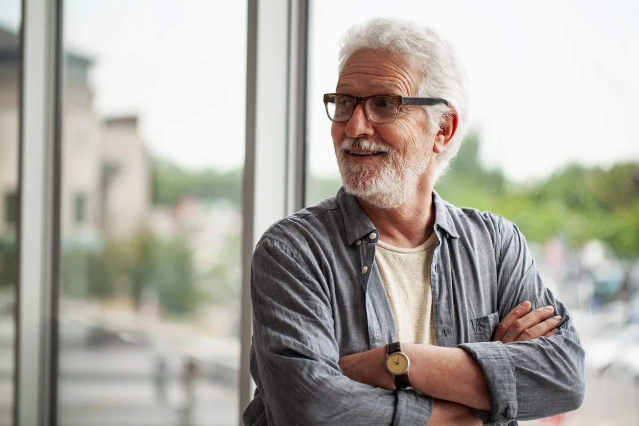 Smiling elderly man with white hair and glasses wearing a blue shirt and watch, standing with arms crossed.