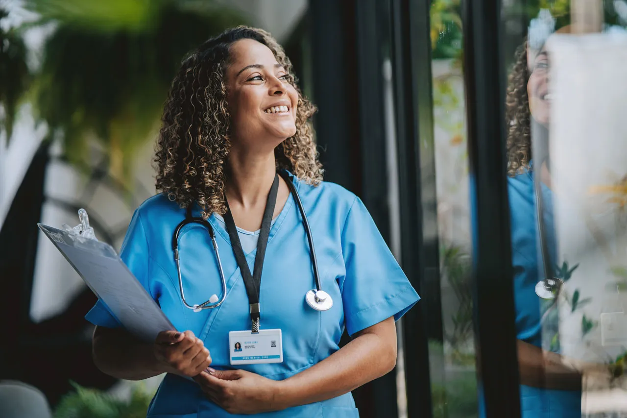Smiling nurse in blue scrubs with stethoscope and clipboard looking out window.