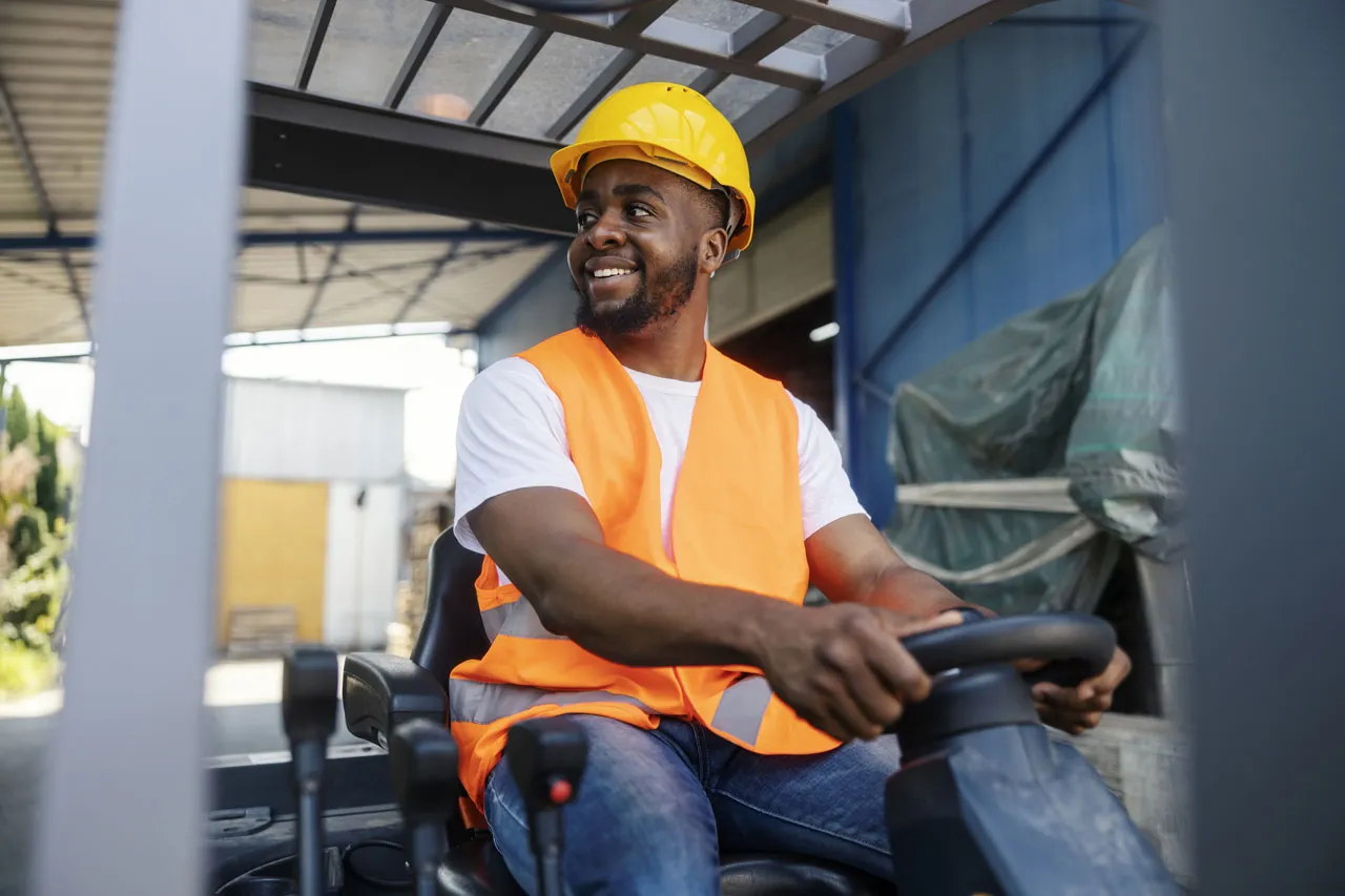 Worker in orange safety vest and yellow helmet driving a forklift in an industrial setting.