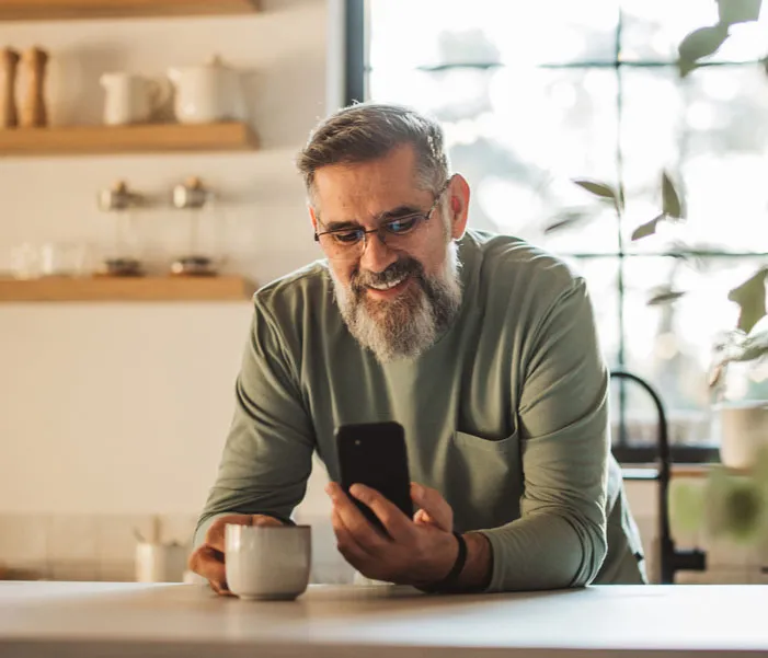Smiling man with glasses and beard holding a smartphone and coffee mug in a bright kitchen.
