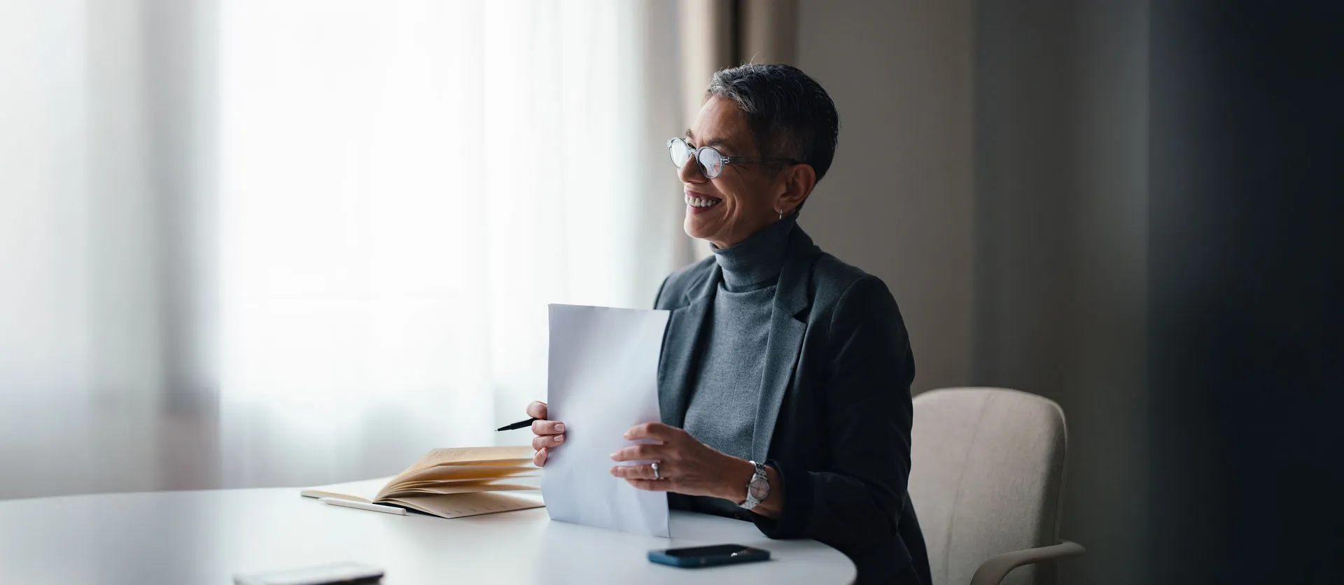 Smiling woman in glasses holding paper with pen, sitting at table with open book and phone.