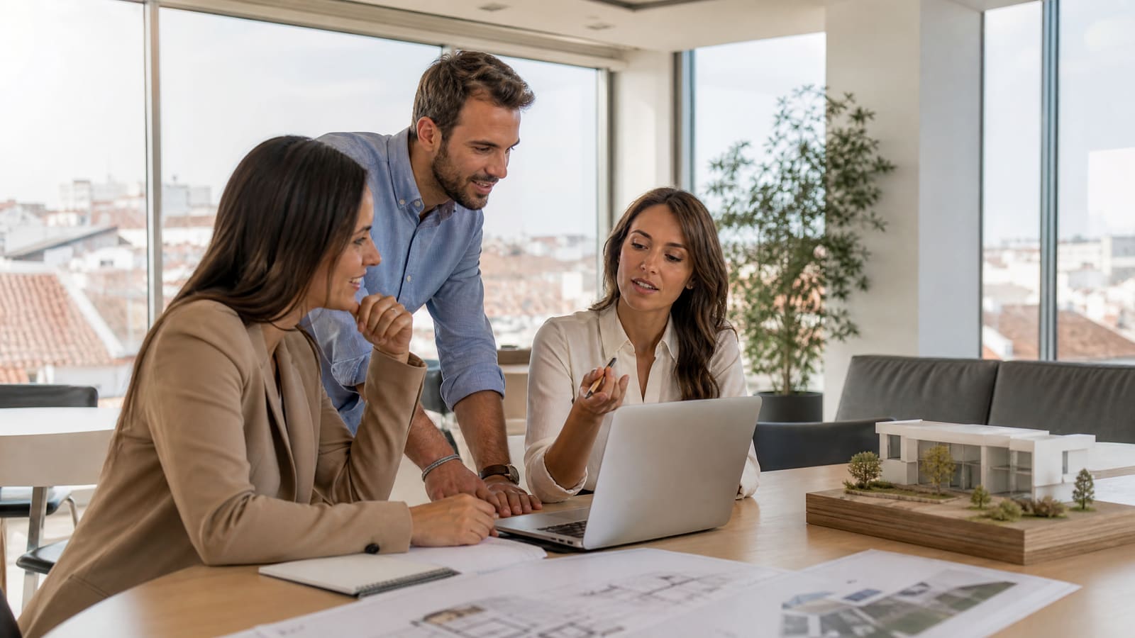 Três colegas consultam crm para imobiliária no portátil, com plantas e maqueta de uma casa na mesa