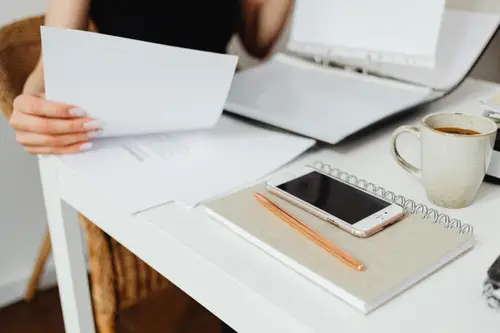 A person seated at a desk holds a stack of white papers while an open laptop sits nearby. A smartphone lies on a notebook with an orange pen, and a cup of coffee is on the right.