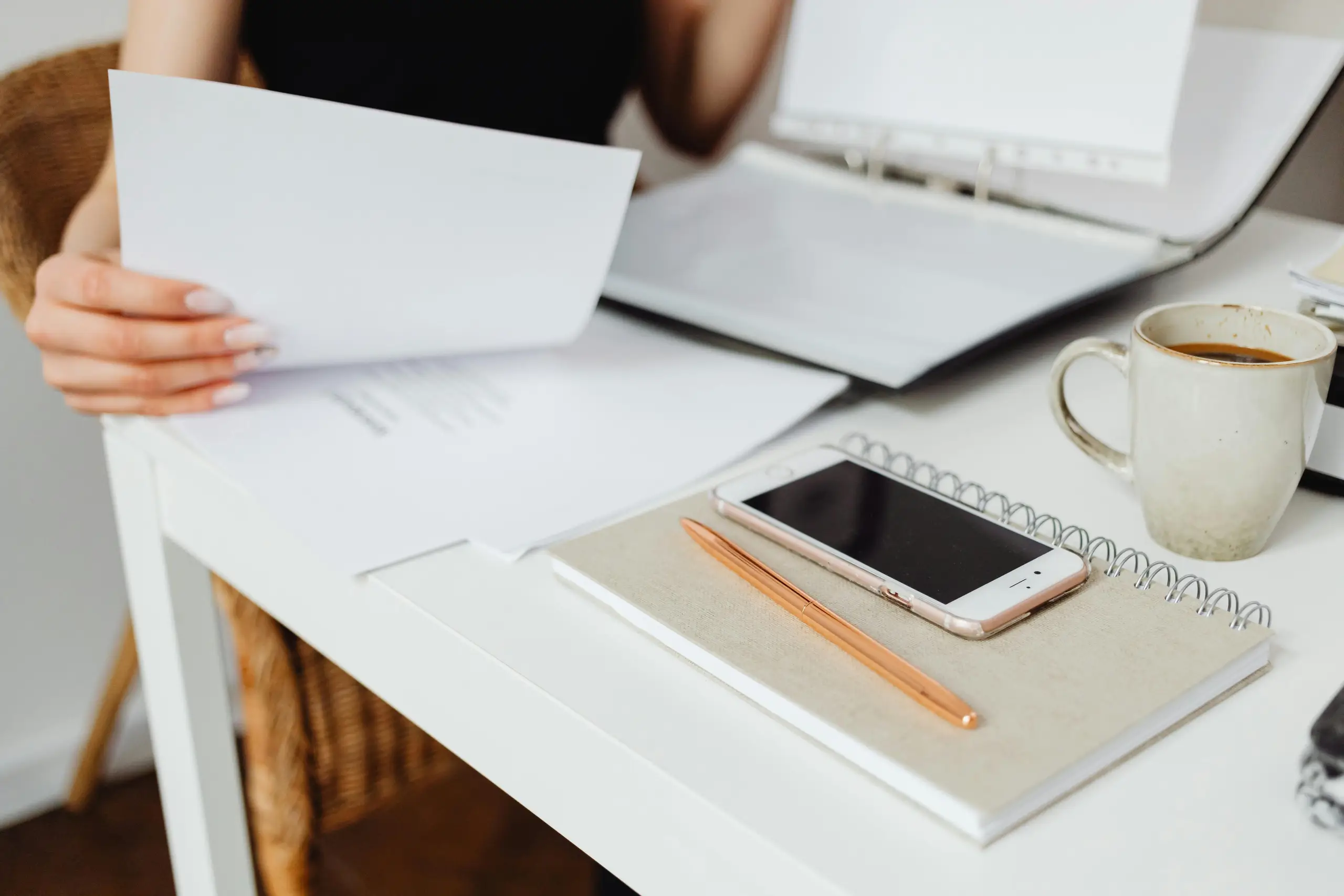A person seated at a desk holds a stack of white papers while an open laptop sits nearby. A smartphone lies on a notebook with an orange pen, and a cup of coffee is on the right.