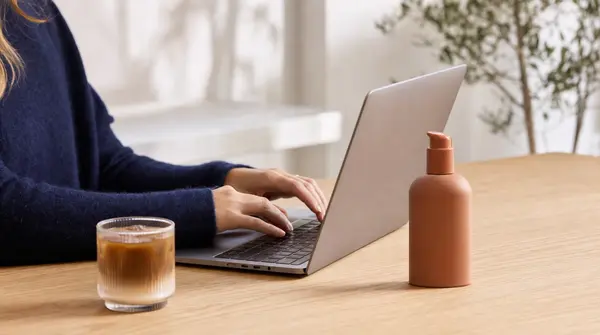 Hands type on a laptop at a wooden table with a beverage and a skincare bottle nearby, suggesting day-to-day operations tied to working capital for ecommerce.