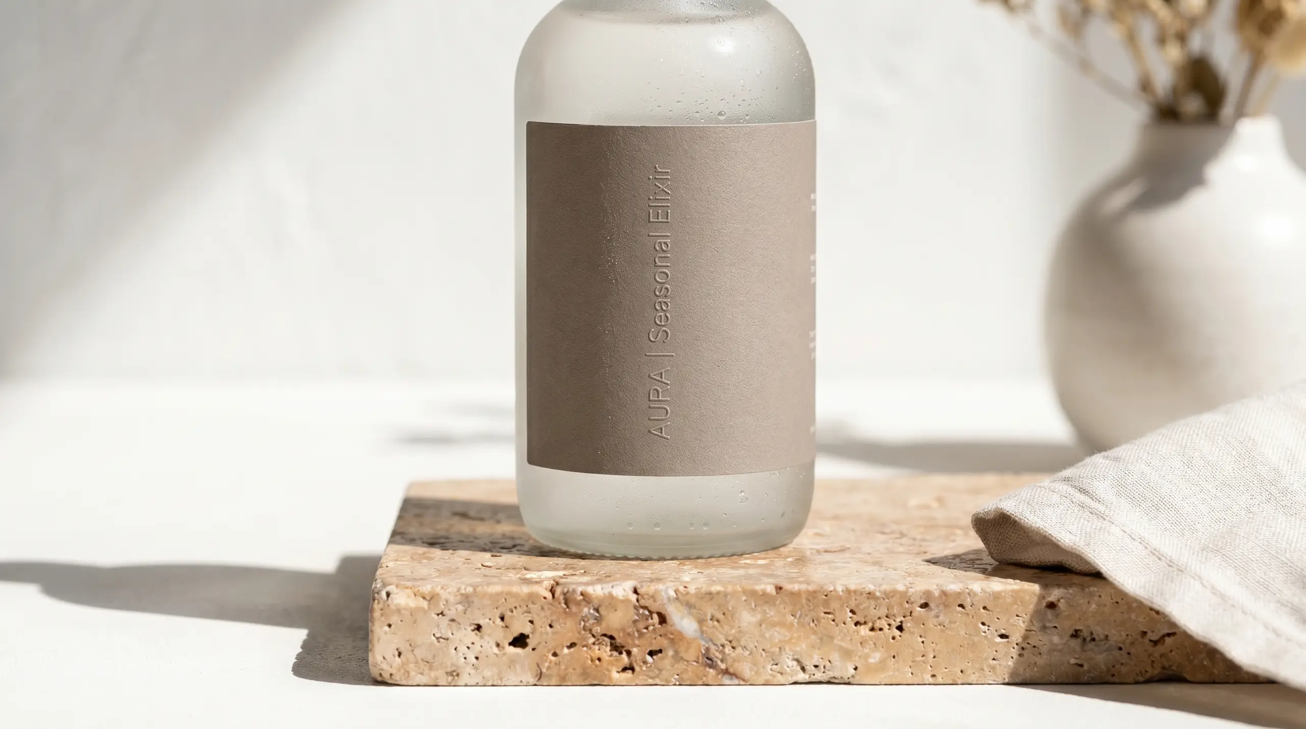 A tall white-and-brown skincare bottle with condensation on it, standing on a rough stone block with a light fabric cloth nearby. The label reads "AUPA | Leaseonaf | Esengai" and "Less.onaf Essentialixir" (vertical text).