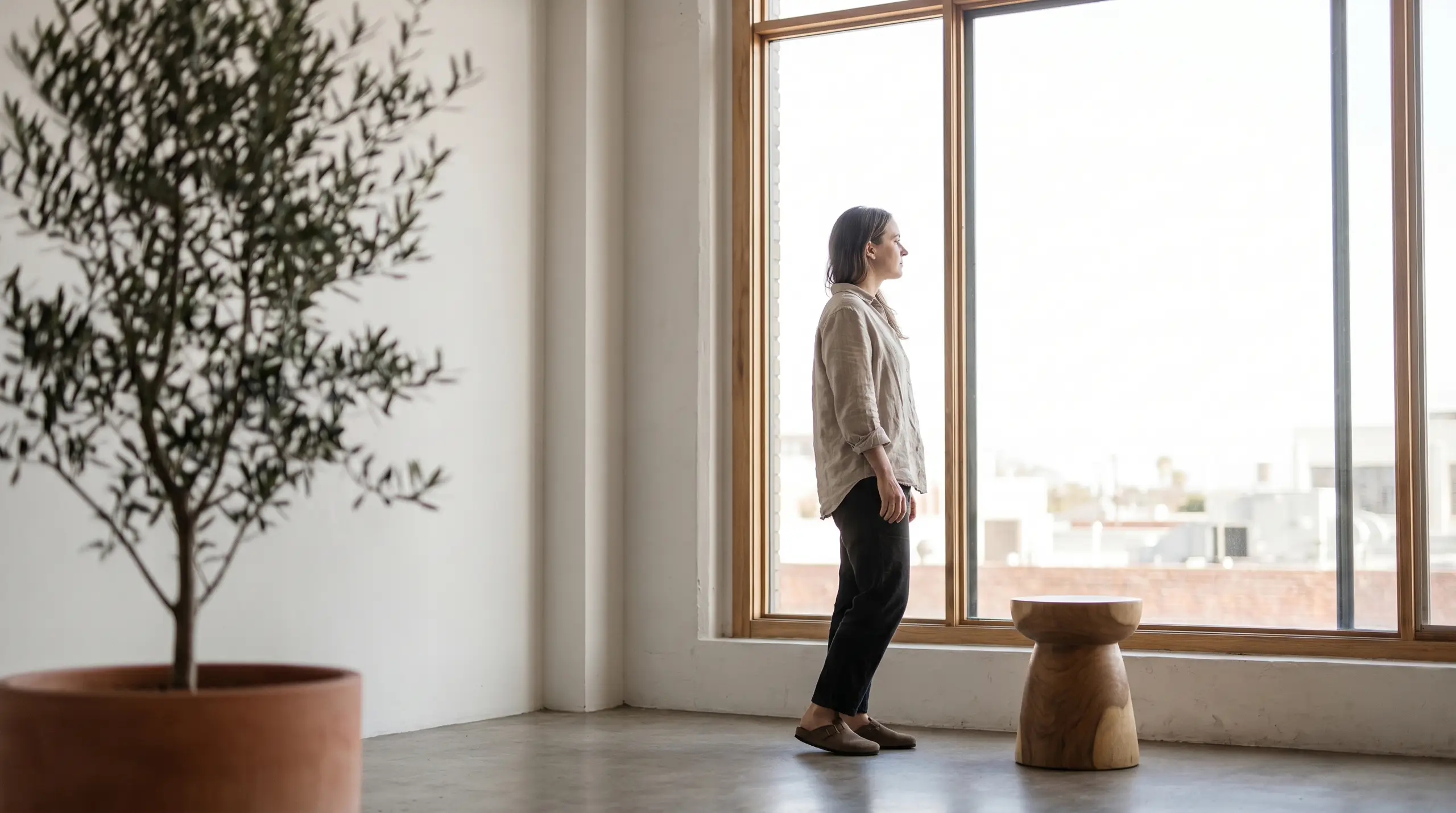 A woman in a light blouse and dark pants stands barefoot near a large wooden-framed window, looking outside. A small tree in a large pot sits to the left, and a wooden stool with a bowl-shaped top rests near the wall on the right.