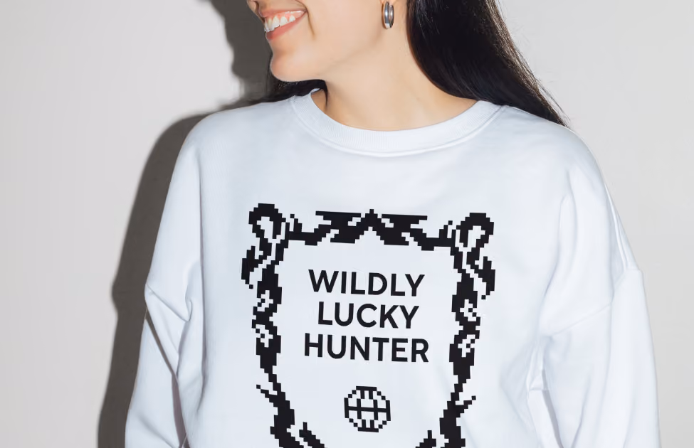 Woman smiling wearing a white sweatshirt with black pixelated frame and text 'WILDLY LUCKY HUNTER'.