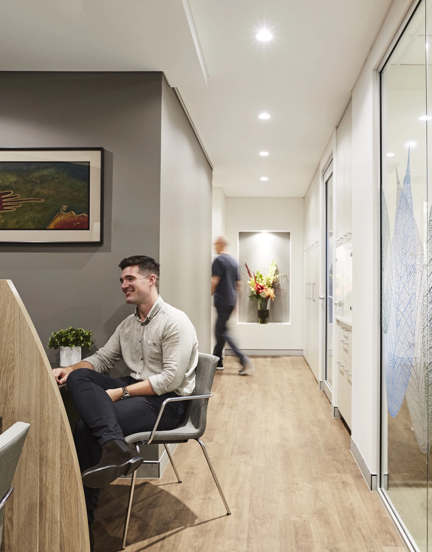 Man sitting and smiling at a wooden desk in a modern office hallway with a blurred person walking in the background.