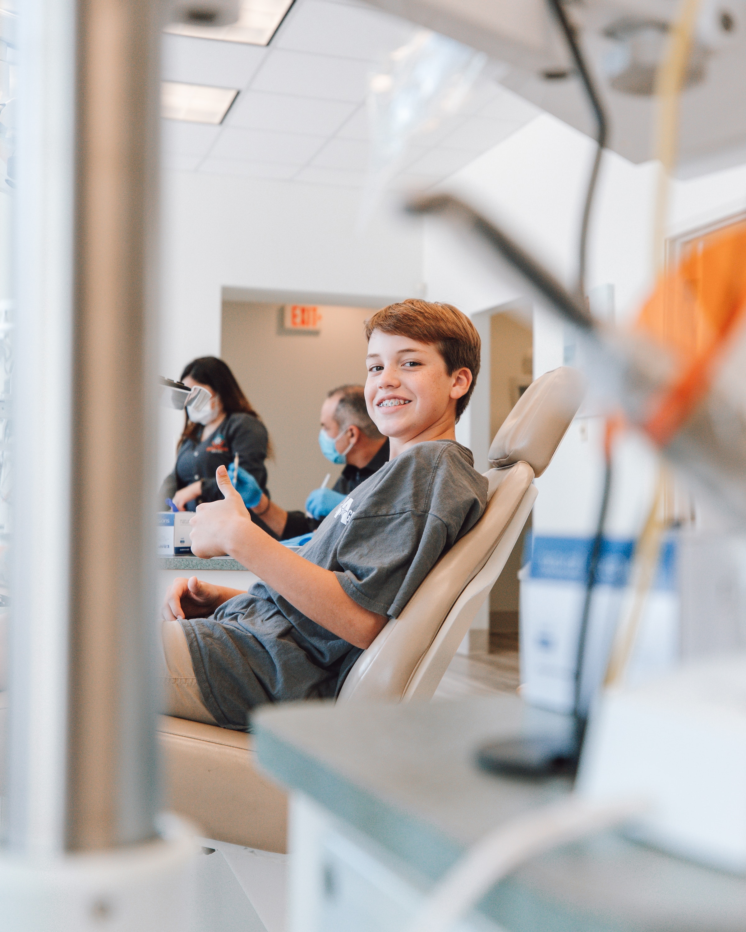 Smiling boy with braces sitting in a dental chair giving a thumbs-up, with two masked dental professionals working in the background.