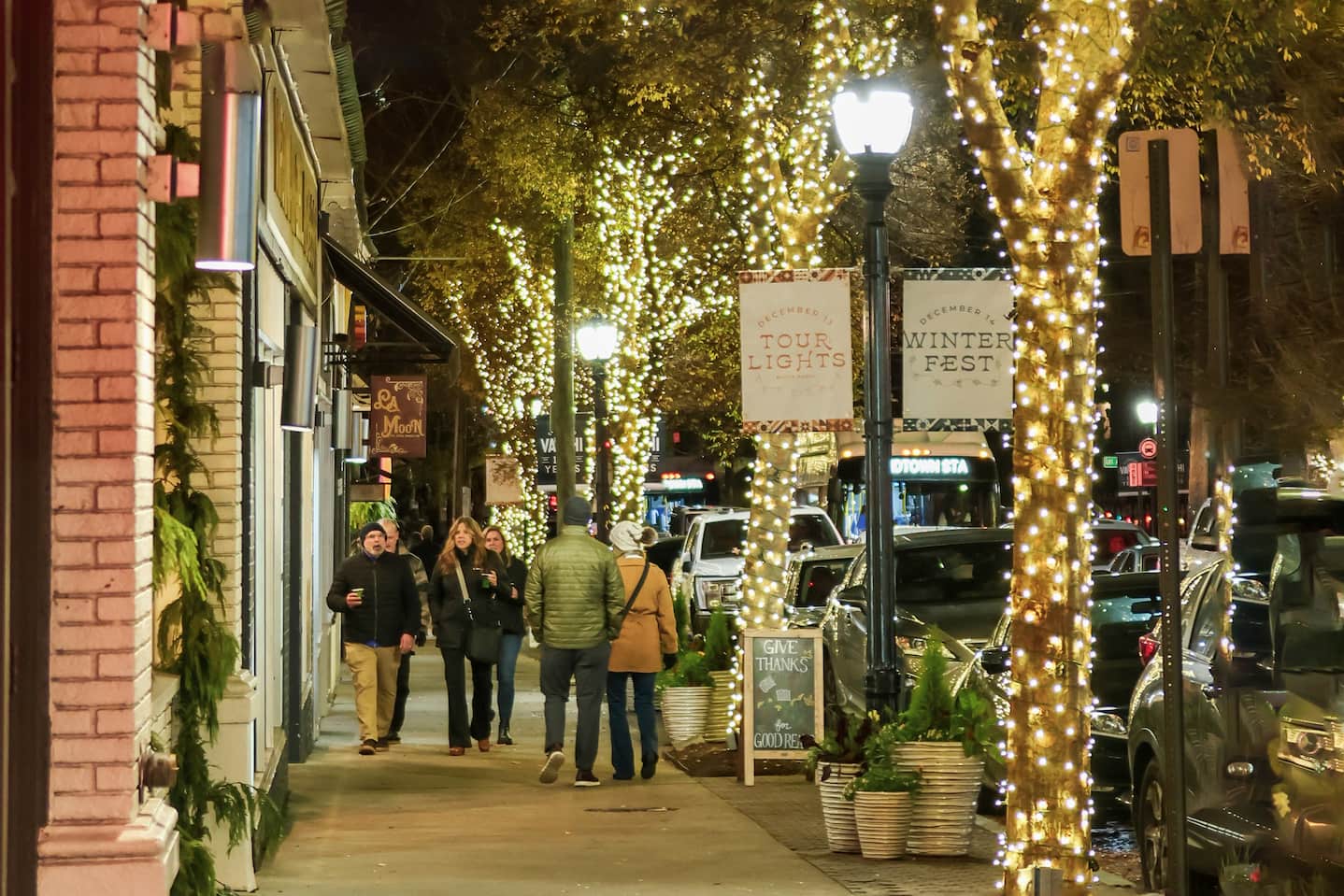 People walking on a city sidewalk at night with trees wrapped in white holiday lights and festive signs advertising 'Tour Lights' and 'Winter Fest'.