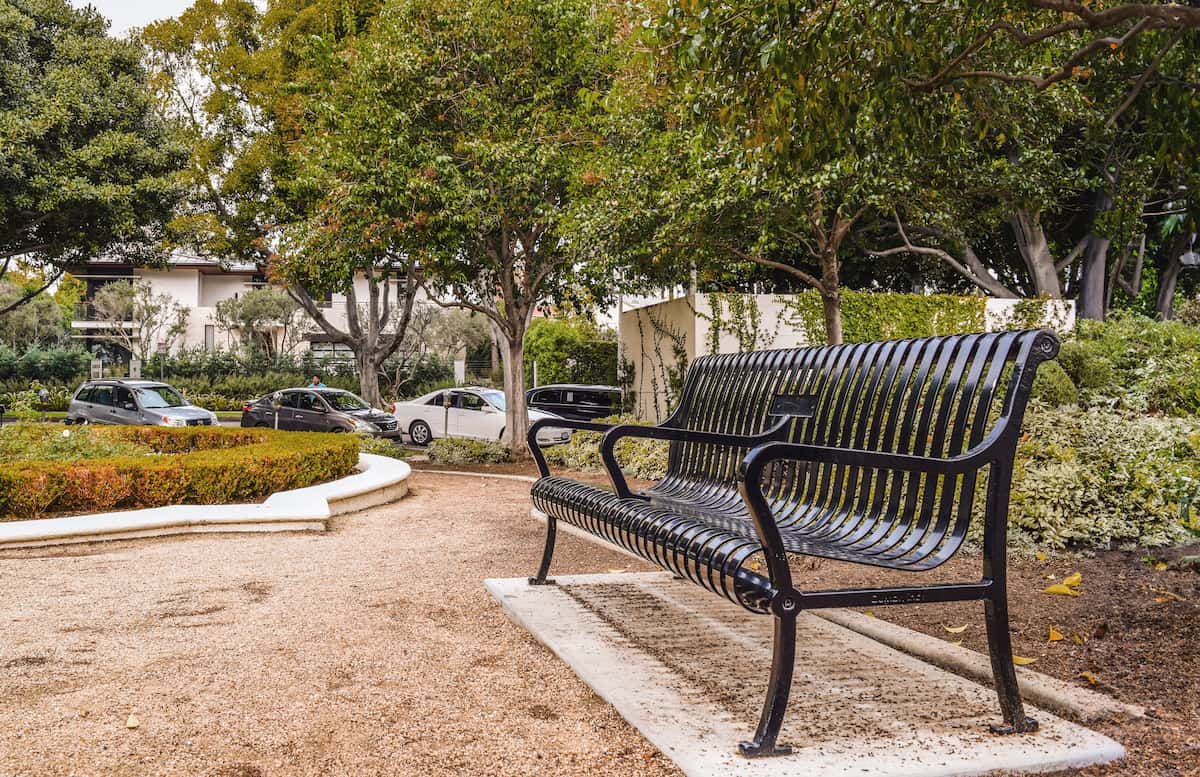 Black metal park bench on gravel path surrounded by green trees and parked cars in the background.