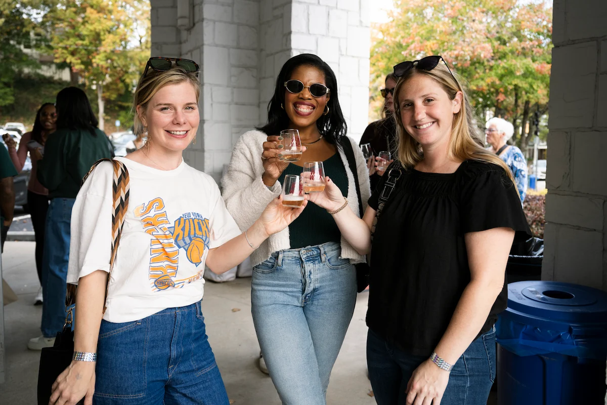 Three women smiling and holding glasses of wine outdoors near stone pillars and autumn trees.