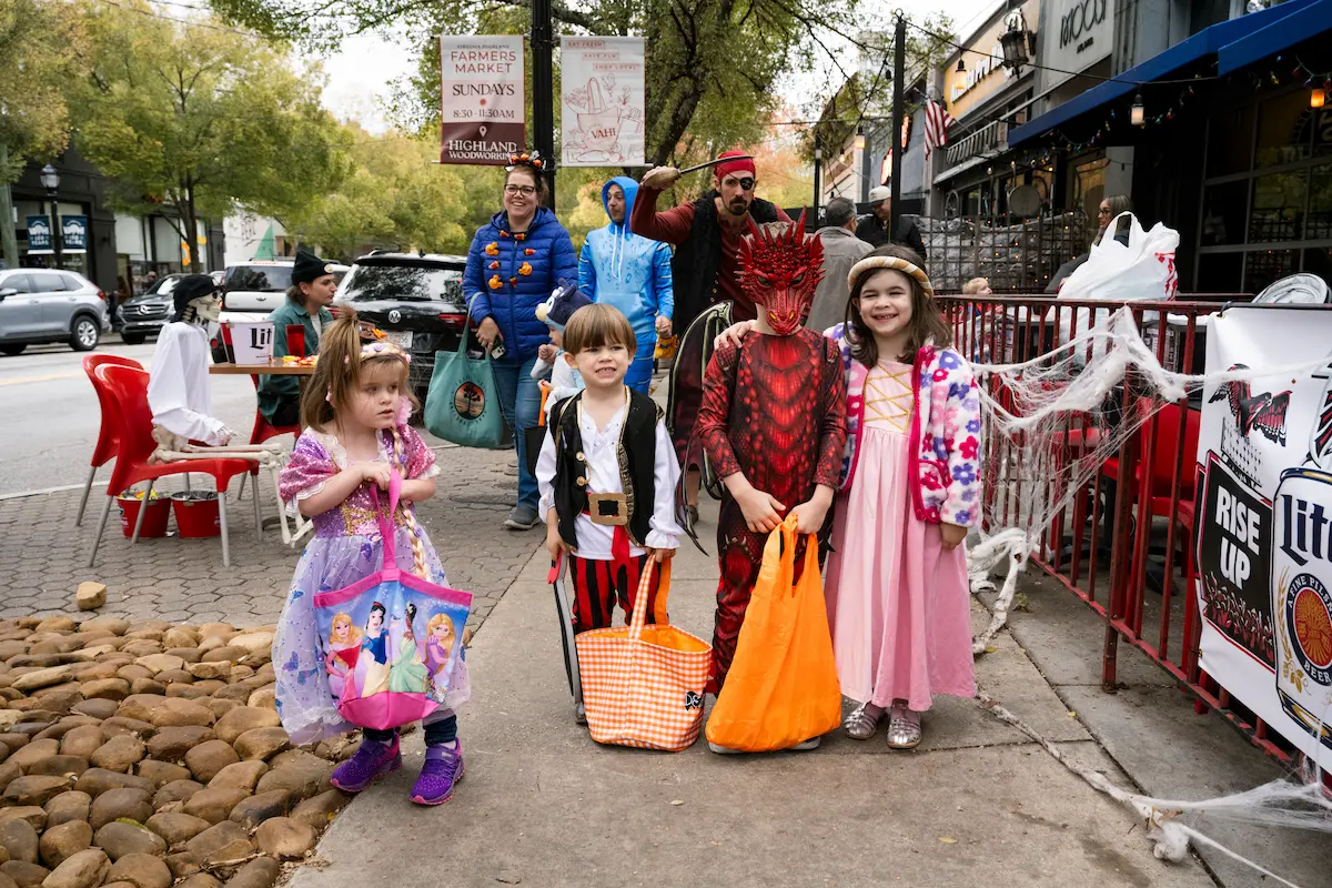 Four children in Halloween costumes holding treat bags on a decorated sidewalk with adults behind them.