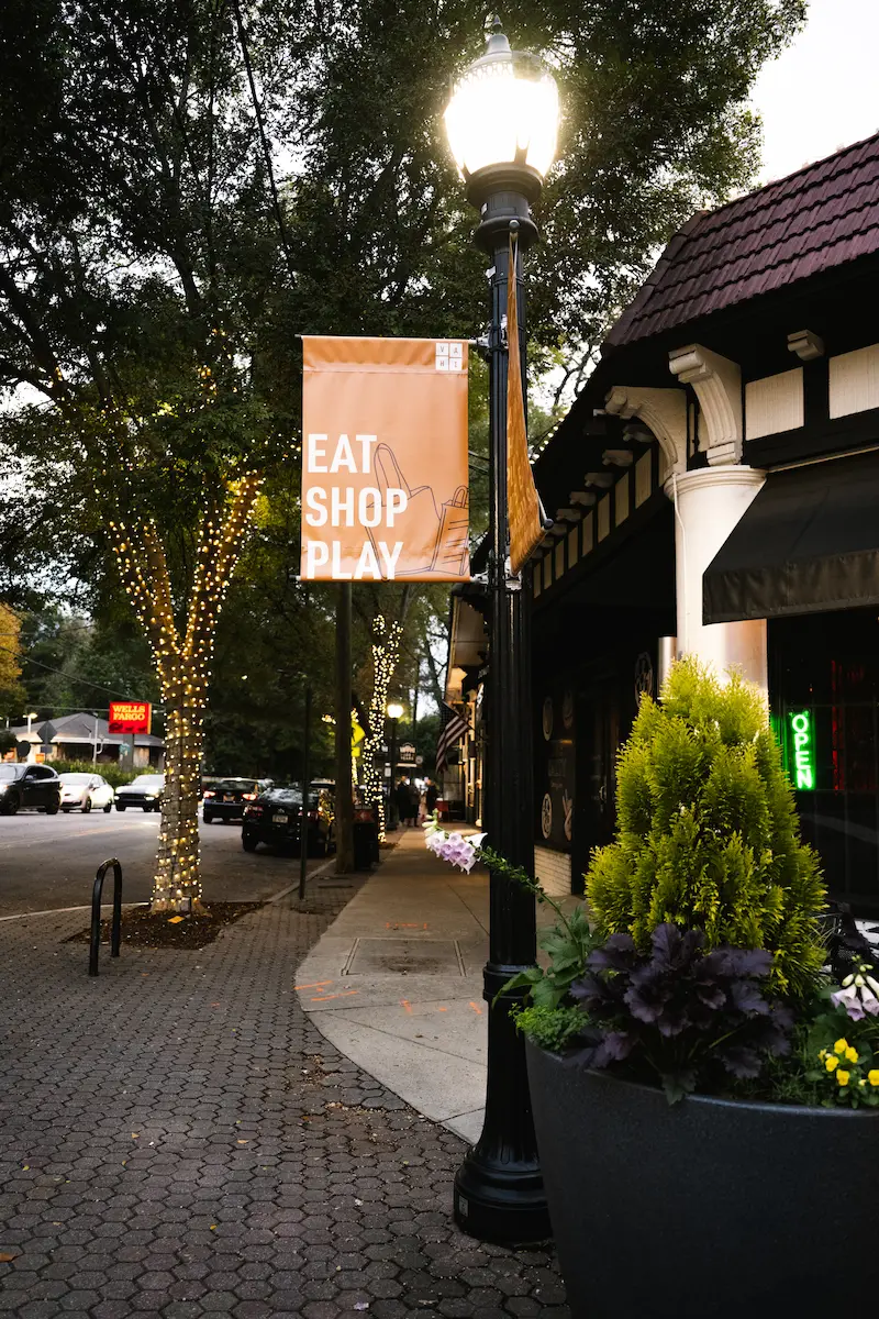Street lamp glowing on a sidewalk with an orange banner reading Eat Shop Play and lit trees nearby.