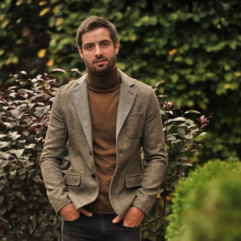 Man with short dark hair and beard wearing a brown turtleneck and gray blazer standing outdoors with hands in pockets.