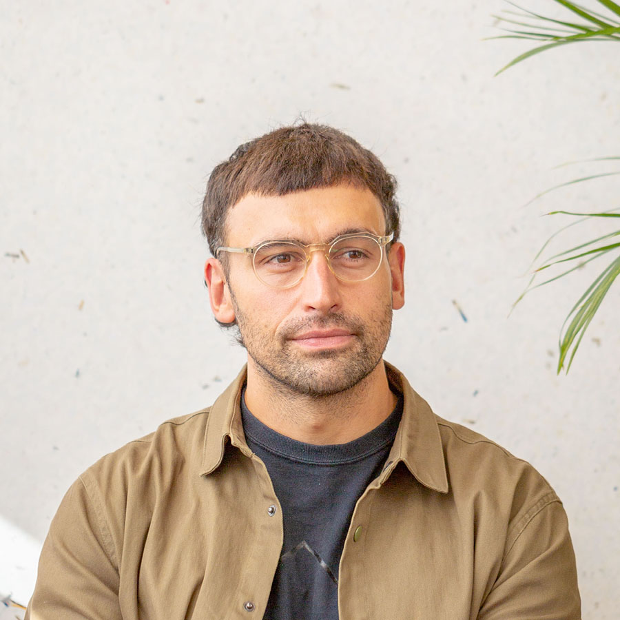 Andy Trewin Hutt wearing glasses and a tan jacket, against a light textured wall.