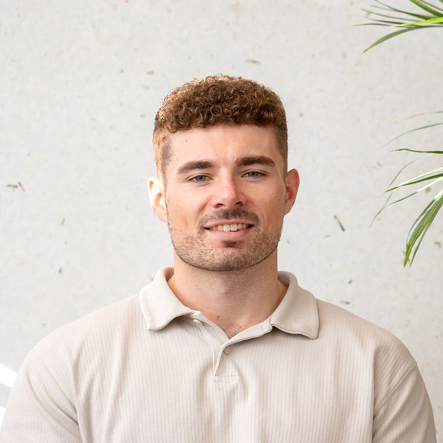 Ben Melvin smiling, wearing a cream ribbed polo, with a plant in the background.