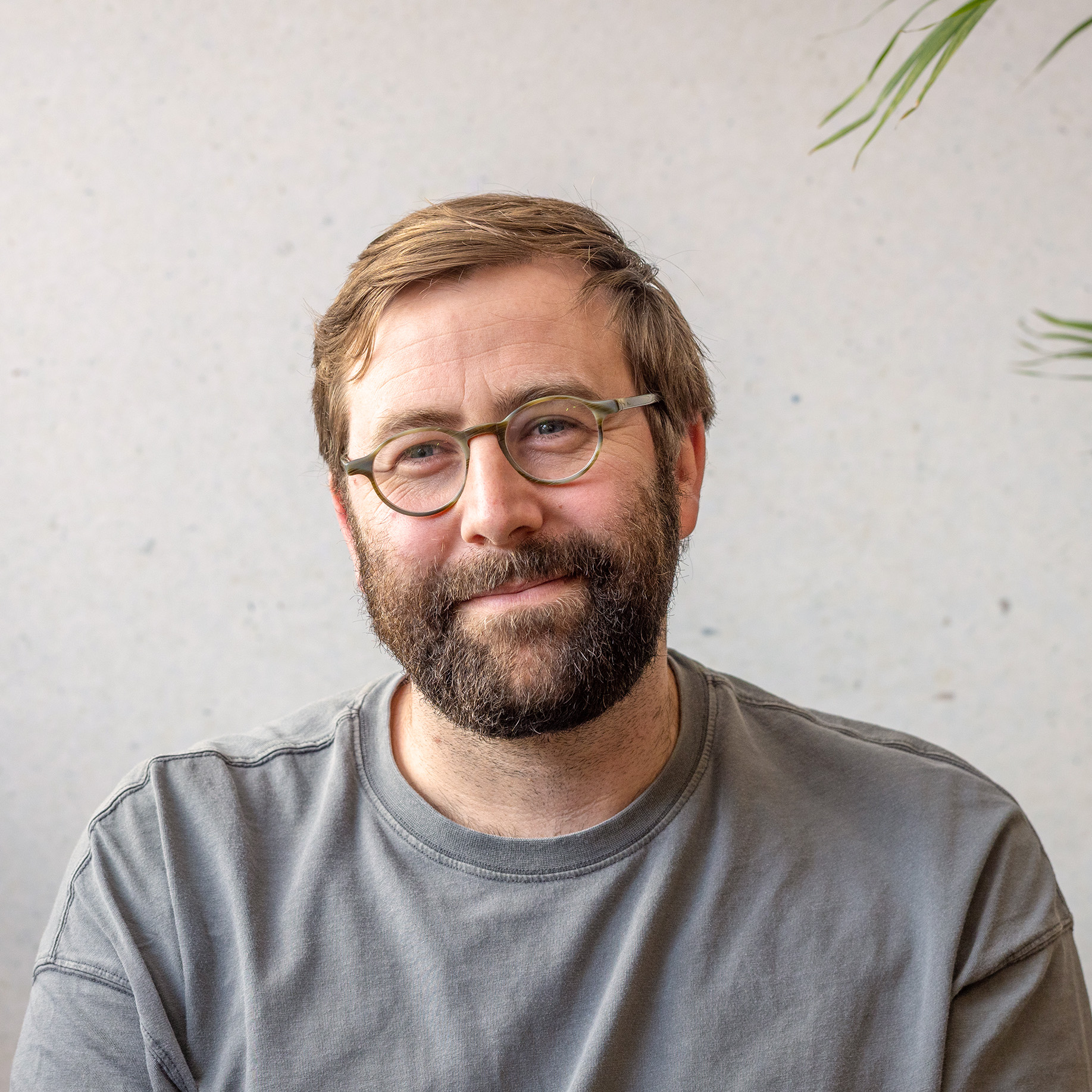 Guy Naor smiling, wearing glasses and a grey t-shirt, with a plant in the background.