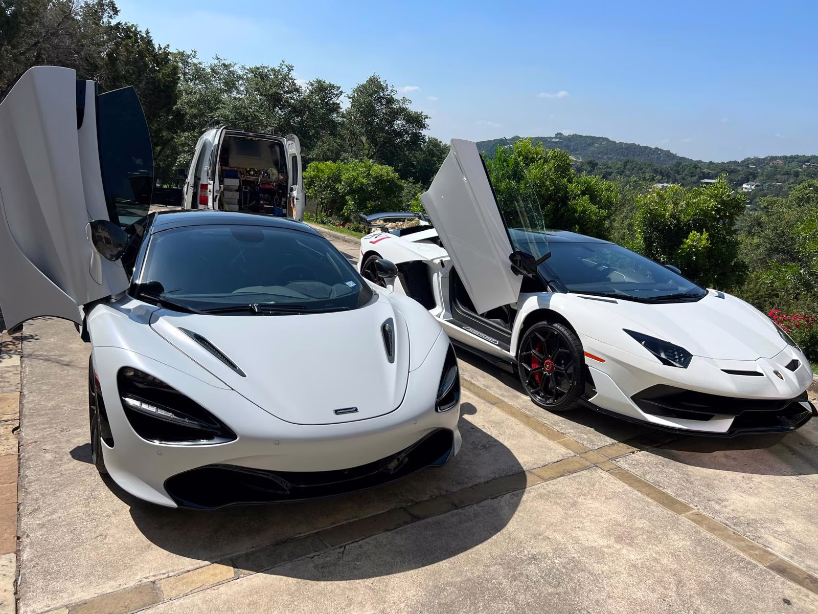 Two white luxury sports cars with raised scissor doors parked outdoors on a concrete driveway with a white van and green trees in the background.