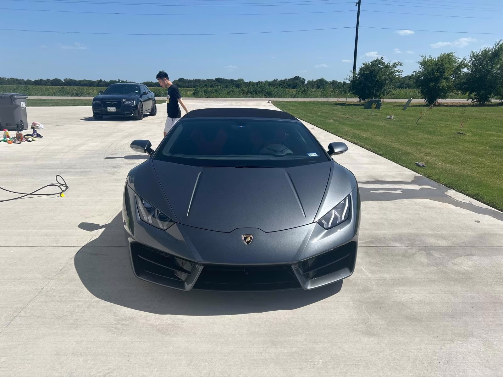 Front view of a gray Lamborghini parked on a concrete driveway with a person walking near a black car in the background.