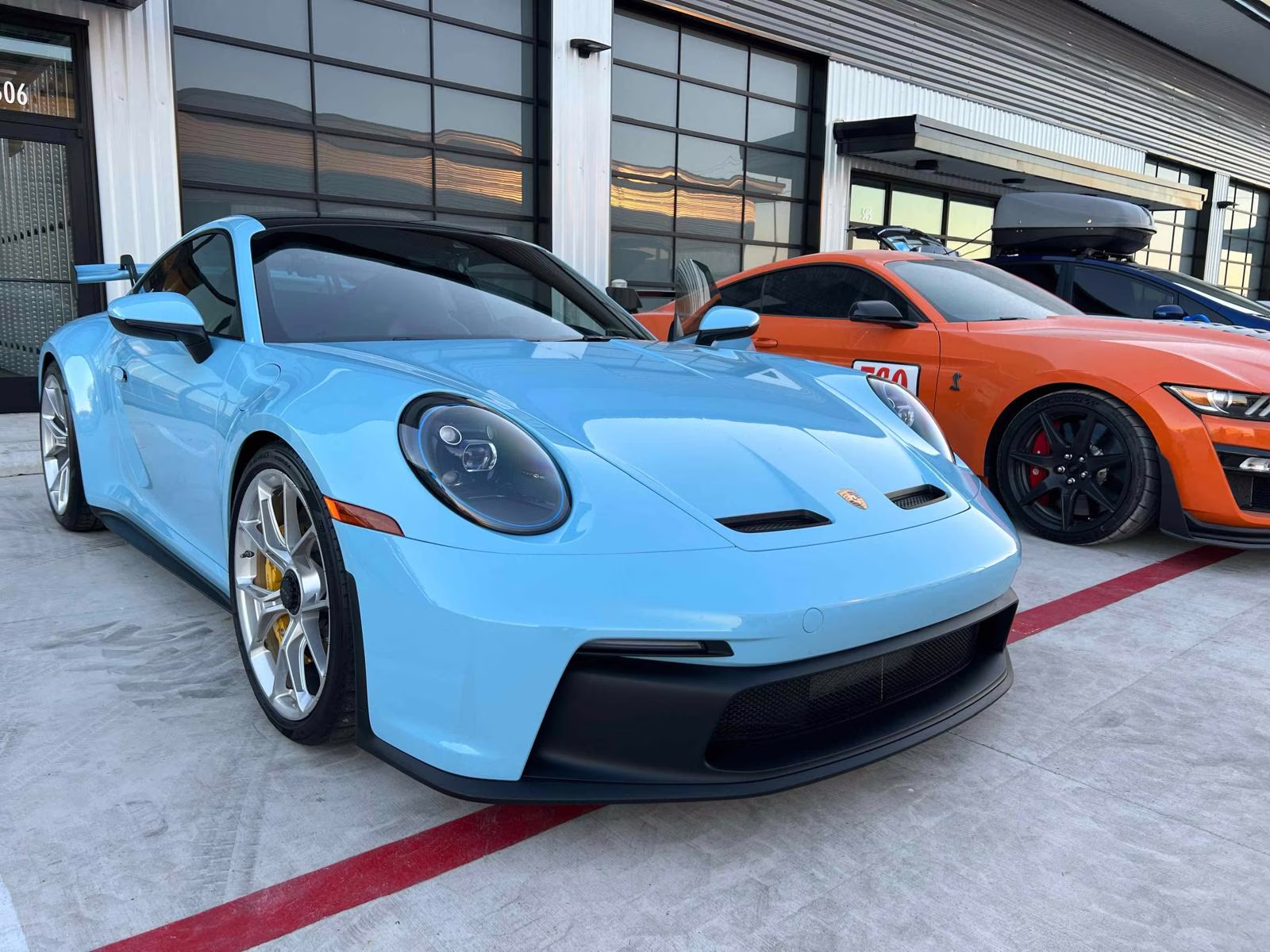 Light blue Porsche sports car parked next to an orange Ford Mustang with black rims in front of a building with large windows.